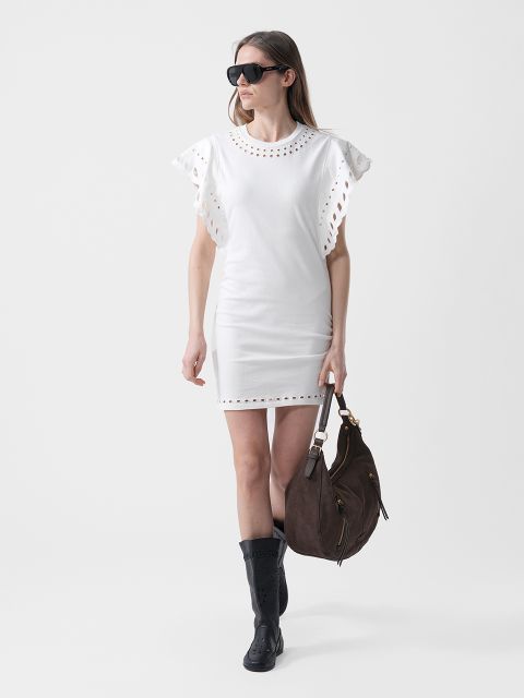 Frau in einem weißen Kleid mit gerafften Ärmeln, schwarzer Sonnenbrille, schwarzen Stiefeln und einer großen braunen Handtasche, die vor einem schlichten Hintergrund geht.