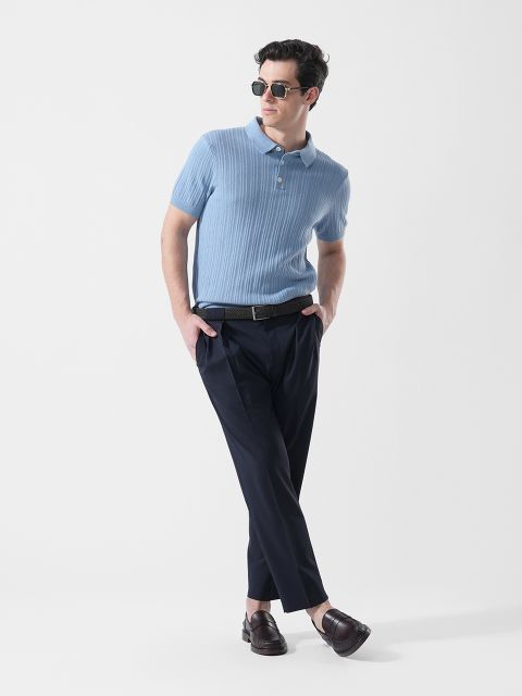 Mann mit Sonnenbrille, der ein hellblaues, kurzärmeliges Polohemd, eine dunkle Hose und braune Slipper trägt und mit einem Fuß vor dem anderen gekreuzt steht.