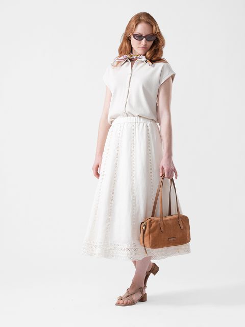 Frau mit Sonnenbrille, weißer Bluse und langem weißen Rock, die eine hellbraune Handtasche hält und beigefarbene Sandalen trägt, steht vor einem weißen Hintergrund.