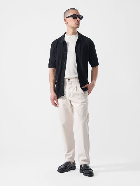 Mann mit Sonnenbrille, der ein schwarzes kurzärmeliges Hemd über einem weißen T-Shirt, eine cremefarbene Hose und schwarze Schuhe trägt und vor einem weißen Hintergrund steht.