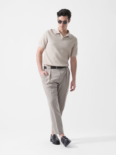 Ein Mann, der eine Sonnenbrille, ein beiges kurzärmeliges Strickhemd, eine hellgraue Hose und schwarze Slipper trägt, steht vor einem schlichten weißen Hintergrund.