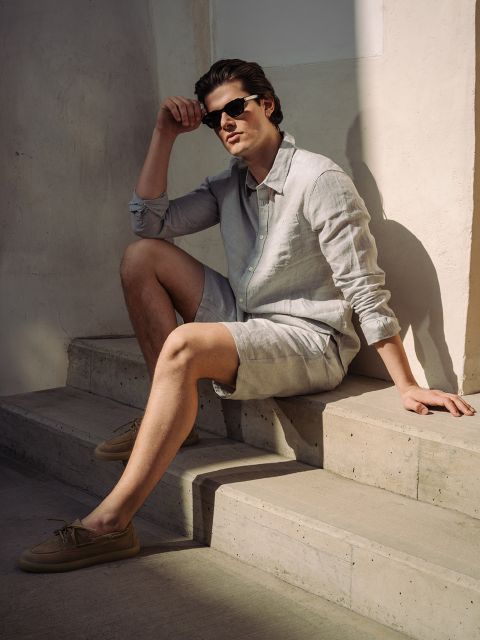 Ein Mann mit Sonnenbrille, hellem Leinenhemd und kurzen Hosen sitzt auf einer Betontreppe im Sonnenlicht und stützt einen Arm auf sein Knie.
