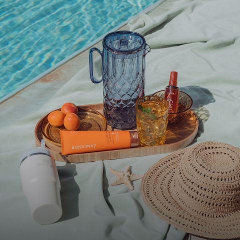 Ein Tablett am Pool mit Glaswaren, Aprikosen, Sonnencreme, einer Sprühflasche, einem Sonnenhut, einem weißen Becher und einem Seestern auf einem hellgrünen Handtuch.