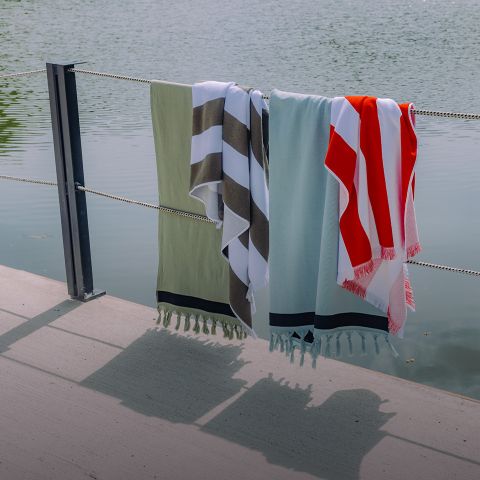 Vier bunte Handtücher hängen über einen Seilzaun am Wasser und werfen Schatten auf die Betonfläche darunter.