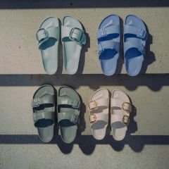 Vier Paar Sandalen in Pastellblau, Pastellgrün, Dunkelgrün und Off-White, die auf einer Betonfläche angeordnet sind und von Schatten durchzogen werden.