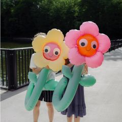 Zwei Personen halten sich große aufblasbare Blumen in Gelb und Rosa vor das Gesicht, während sie auf einem sonnigen Weg in der Nähe eines Geländers und von Grünpflanzen stehen.