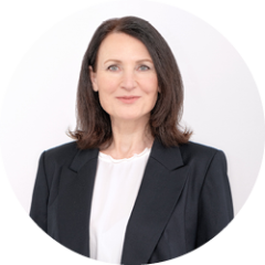 Frau mit schulterlangen braunen Haaren, die einen schwarzen Blazer und eine weiße Bluse trägt, lächelt sanft vor einem schlichten hellen Hintergrund.