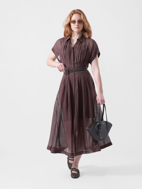 Eine Frau in einem durchsichtigen braunen Kleid mit Gürtel, Sonnenbrille und Sandalen, die eine schwarze Handtasche trägt, geht selbstbewusst vor einem schlichten weißen Hintergrund.