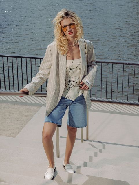 Eine Frau mit lockigem blondem Haar, die eine Sonnenbrille, einen gestreiften Blazer, ein geblümtes Oberteil, Jeansshorts und Slipper trägt, steht an einem Geländer am Wasser.