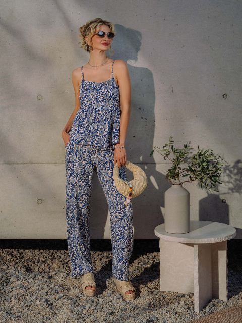 Eine Frau mit Sonnenbrille und geblümtem blauen Outfit steht an einer Betonwand und hält einen Strohhut neben einer Vase mit Olivenzweigen auf einem kleinen Tisch.