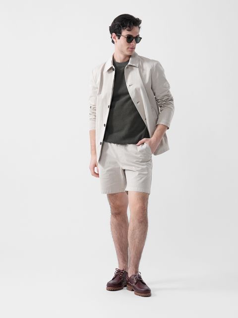 Ein Mann mit Sonnenbrille, beiger Jacke, olivgrünem Hemd, beigen Shorts und braunen Schuhen steht vor einem schlichten weißen Hintergrund.