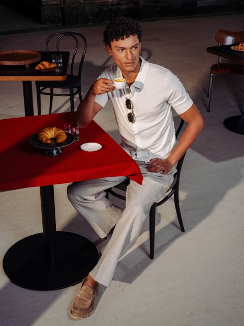 Ein Mann in weißem Hemd und heller Hose sitzt an einem Cafétisch mit Croissants, nippt an einem Espresso und hat eine Sonnenbrille auf dem Hemd.