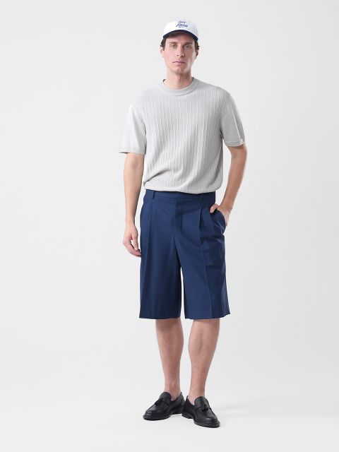 Ein Mann, der eine weiße Mütze, einen hellgrauen kurzärmeligen Pullover, marineblaue knielange Shorts und schwarze Slipper trägt, steht vor einem schlichten weißen Hintergrund.