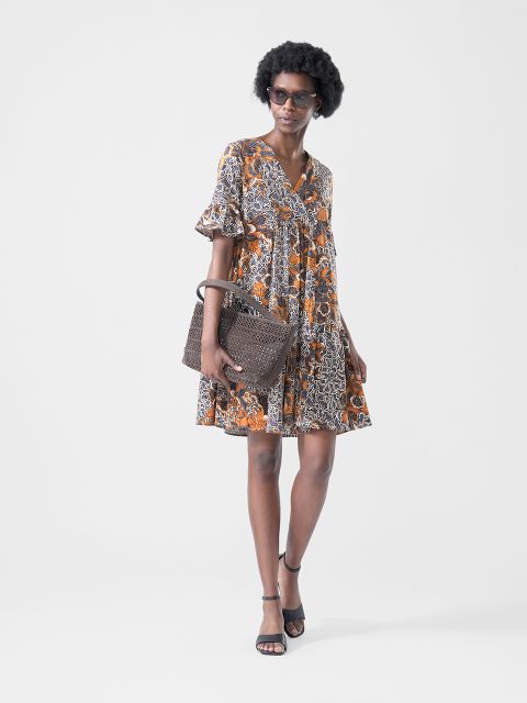 Frau trägt ein kurzes, gemustertes Kleid mit orangefarbenen und schwarzen Tönen, eine Sonnenbrille, schwarze Sandalen und hält eine braune, strukturierte Handtasche.