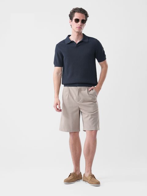 Mann mit Sonnenbrille, marineblauem, kurzärmeligem Polohemd, beigen, knielangen Shorts und braunen Bootsschuhen vor einem schlichten weißen Hintergrund.