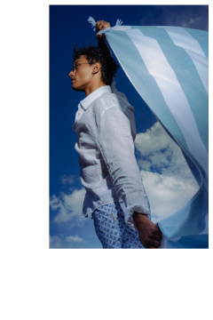 Eine Person in einem weißen Hemd und gemusterten Shorts hält ein blau-weiß gestreiftes Tuch vor einen blauen Himmel mit Wolken.