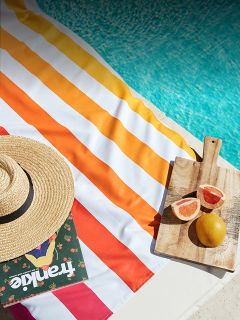 Ein gestreiftes Strandtuch, ein Strohhut, eine Zeitschrift und Obst auf einem Schneidebrett liegen am Rand eines Pools mit türkisfarbenem Wasser.