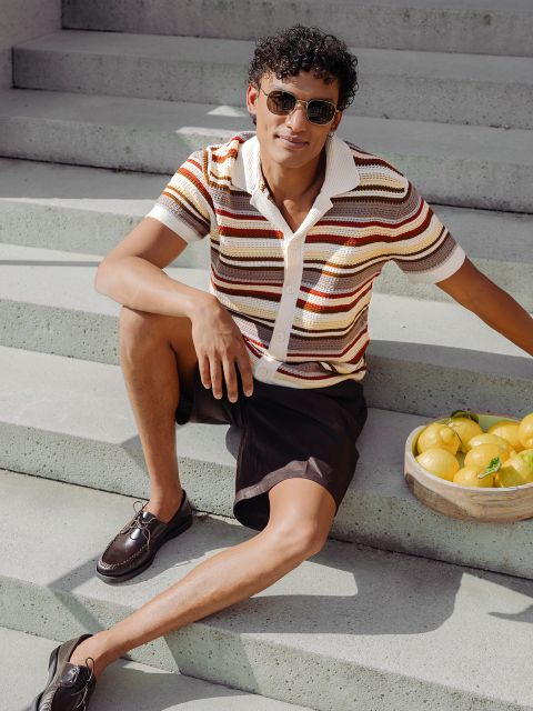 Ein Mann mit Sonnenbrille und gestreiftem Hemd sitzt auf einer Freitreppe neben einem Korb mit Zitronen und lächelt lässig in das Sonnenlicht.