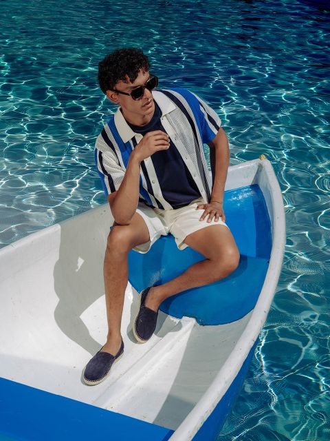 Ein Mann mit Sonnenbrille und gestreiftem Hemd sitzt auf einem kleinen weiß-blauen Boot in einem Pool mit klarem, glitzerndem Wasser.