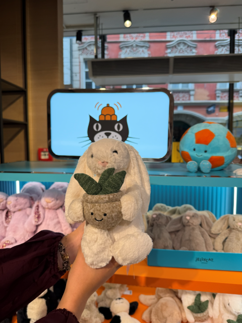 Person holding up a white plush rabbit wearing a small plant pot around its neck in a toy store display, with other stuffed animals visible in the background.