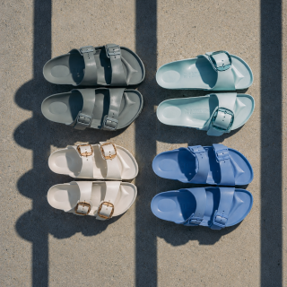 Vier Paar Sandalen in Grau, Hellblau, Creme und Blau sind auf Beton zu einem Quadrat angeordnet, über das Schatten fallen.