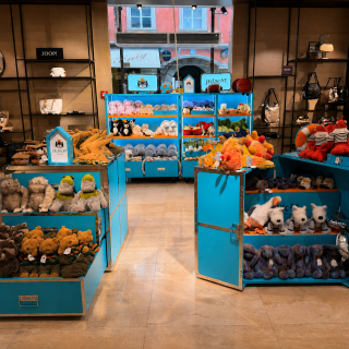 Ein hell beleuchtetes Geschäft mit blauen Regalen, in denen verschiedene Plüschtiere ausgestellt sind, darunter Hunde, Dinosaurier und Bären. Im Hintergrund hängen Handtaschen an Wandregalen.