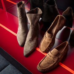 Zwei Paar braune Wildlederschuhe – Stiefeletten und Slipper –, die auf einem glänzenden roten Regal ausgestellt sind, in dem sich ihre Spiegelbilder widerspiegeln.