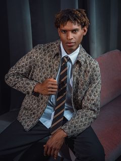 Ein Mann in einer gemusterten Jacke, einem Hemd und einer gestreiften Krawatte sitzt auf einem roten Sofa vor einem dunklen Vorhang und blickt selbstbewusst in die Kamera.