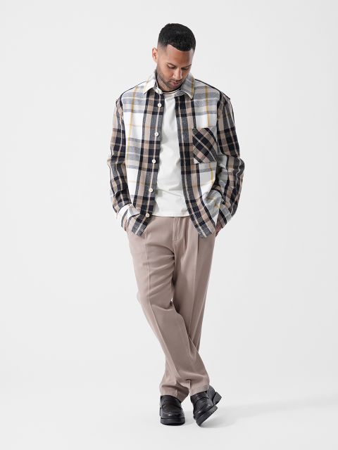 Ein Mann, der ein kariertes Hemd über einem weißen T-Shirt, eine beige Hose und schwarze Schuhe trägt, steht mit den Händen in den Taschen da und blickt vor einem schlichten weißen Hintergrund nach unten.