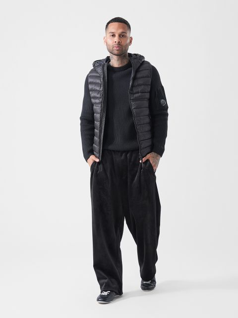 Ein Mann, der eine schwarze Daunenweste mit Kapuze, einen schwarzen Pullover, eine schwarze Hose mit weitem Bein und schwarze Schuhe trägt, steht vor einem schlichten weißen Hintergrund.