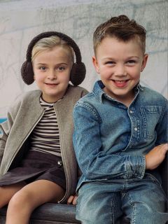 Zwei kleine Kinder sitzen nebeneinander, ein Mädchen mit Ohrenschützern und einer Jacke und ein Junge in einem Jeanshemd und Jeans; beide lächeln in die Kamera.