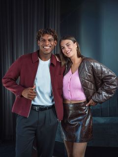 Ein lächelnder Mann in einer burgunderroten Jacke steht neben einer Frau in einem rosa Oberteil und einem braunen Rock; beide posieren in einem Innenraum vor einem dunklen Hintergrund.