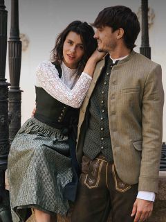 Eine Frau und ein Mann in traditioneller bayerischer Tracht sitzen zusammen im Freien, lächeln und schauen sich an.