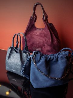 Drei stilvolle Handtaschen – eine aus burgunderrotem Wildleder, eine aus marineblauem Leder und eine aus marineblauem Wildleder mit Kettenriemen – sind vor einem rotbraunen Hintergrund ausgestellt.