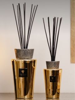 Zwei goldfarbene Vasen mit gemusterten Oberteilen, in denen hohe schwarze Diffusorstäbchen stecken, stehen auf einer Ablage vor einer neutralen Wand.