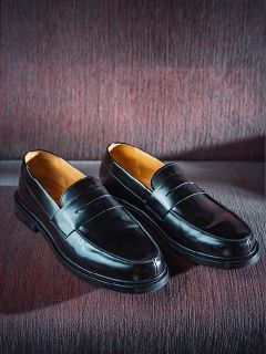 Ein Paar glänzende schwarze Leder-Loafer, die nebeneinander auf einer strukturierten, weinroten Stoffunterlage liegen.
