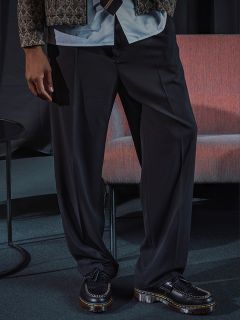 Eine Person, die eine weite schwarze Hose, eine gemusterte Jacke und schwarze Slipper trägt, steht neben einem rosa Stuhl und einem kleinen runden Tisch.