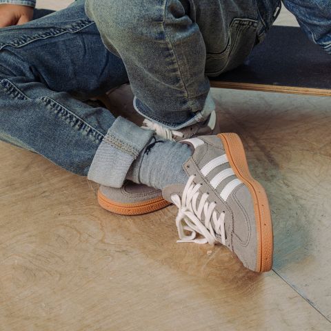 Eine Person in blauen Jeans und grauen Turnschuhen mit weißen Streifen sitzt auf einem Skateboard auf einem Holzboden, die Beine an den Knöcheln übereinandergeschlagen.