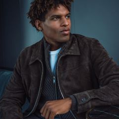 Ein Mann, der eine dunkelbraune Wildlederjacke, einen grauen Pullover und ein weißes Hemd trägt, sitzt in einem Raum und blickt mit nachdenklichem Gesichtsausdruck zur Seite.