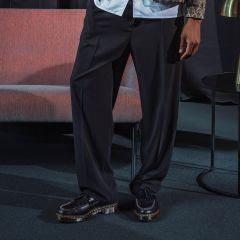 Eine Person steht in einem Innenraum und trägt eine schwarze Hose mit weitem Bein und schwarze Plateauschuhe; neben ihr stehen ein rosa Sofa und ein kleiner runder Tisch.