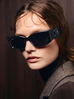 Eine Frau mit langen braunen Haaren, die eine große schwarze Sonnenbrille und einen braunen Mantel trägt und vor einem dunklen Holzhintergrund posiert.