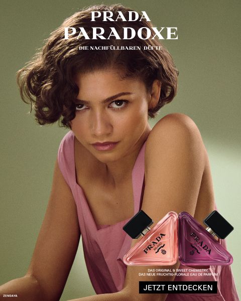 Eine Frau in einem rosa Kleid posiert hinter zwei „Prada Paradoxe“-Parfümflakons vor einem grünen Hintergrund. Der Text wirbt auf Deutsch für den Duft.