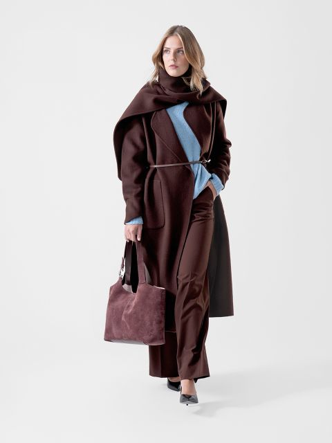 Eine Frau in einem dunkelbraunen Mantel und einer dunkelbraunen Hose sowie einem blauen Pullover, die eine dazu passende braune Tasche in der Hand hält und selbstbewusst vor einem schlichten weißen Hintergrund geht.