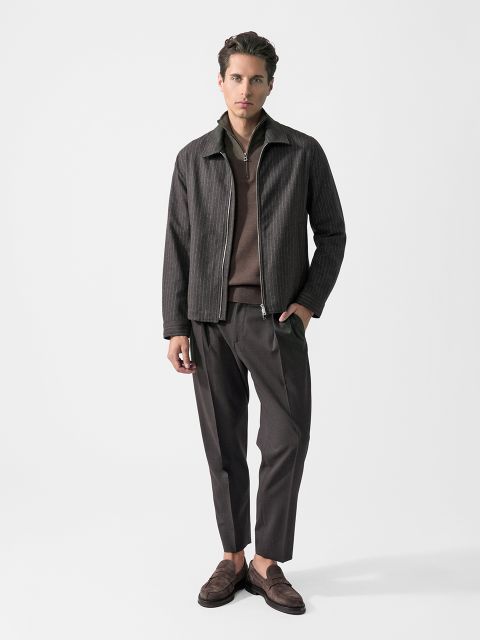 Ein Mann, der eine dunkel gestreifte Jacke, einen braunen Pullover, eine dunkle Hose und braune Slipper trägt, steht vor einem schlichten weißen Hintergrund.