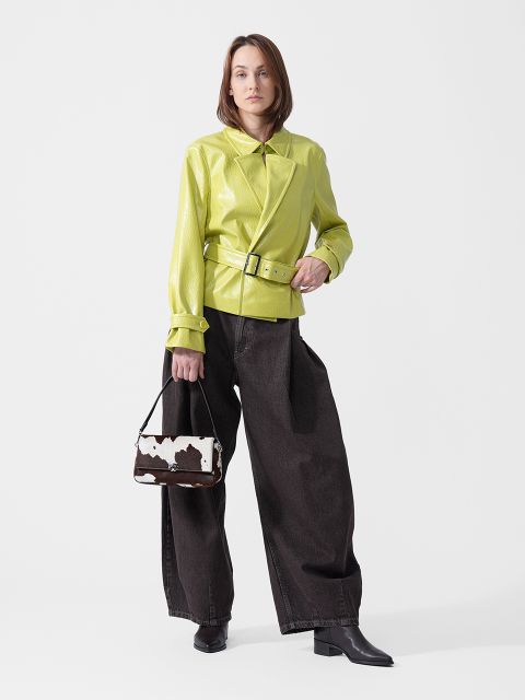 Eine Frau steht da in einer limettengrünen Jacke mit Gürtel, einer dunkelblauen Jeans mit weitem Bein und schwarzen Stiefeln und hält eine kleine Handtasche mit Kuhmuster in der Hand.