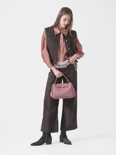 Eine Frau in brauner Weste und Hose, rosa Bluse und schwarzen Stiefeln hält eine rosa Handtasche in der Hand und steht vor einem schlichten weißen Hintergrund.
