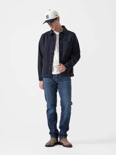 Ein Mann, der eine marineblaue Jacke, ein weißes T-Shirt, blaue Jeans, graue Stiefel und eine weiße NY-Baseballkappe trägt, steht vor einem schlichten weißen Hintergrund.