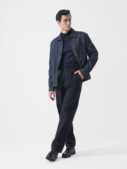 Ein Mann, der eine dunkle Jeansjacke, einen marineblauen Rollkragenpullover, eine schwarze Hose und schwarze Schuhe trägt, steht vor einem schlichten weißen Hintergrund und blickt nach links.