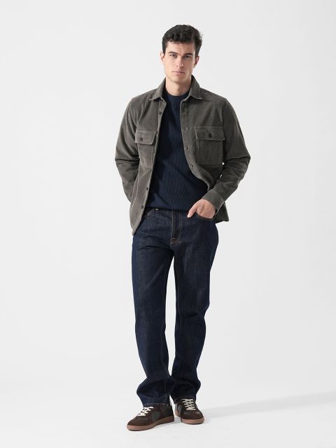 Ein Mann in einer grauen Jacke, dunkelblauen Jeans und braunen Sneakers steht vor einem schlichten weißen Hintergrund und hat die Hände in den Taschen.
