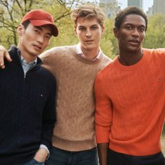 Three men posing in a park, wearing sweaters (navy, beige, orange); the left man has his arm around the middle man, with a city skyline in the background.